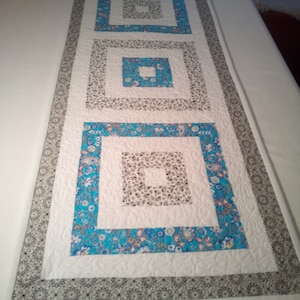 Könnte beinhalten: Ein weißer Tischläufer mit einem blauen und weißen Blumenmuster. Der Läufer hat drei Schichten von Quadraten, jede mit einem anderen Muster. Die äußere Schicht ist ein graues und weißes Muster.