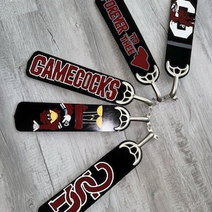 May include: A collection of black ceiling fan blades with various designs. One blade reads "GAMECOCKS" in maroon and white. Other blades feature the text "FOREVER TO THEE", the letters "USC", and a mascot illustration. Each blade has a silver metal attachment.