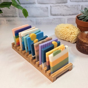 May include: A wooden soap holder displaying a variety of colorful, layered soap bars. The soaps feature shades of blue, green, yellow, orange, and purple. The holder is made of light brown wood and is labeled "SALT TECH".