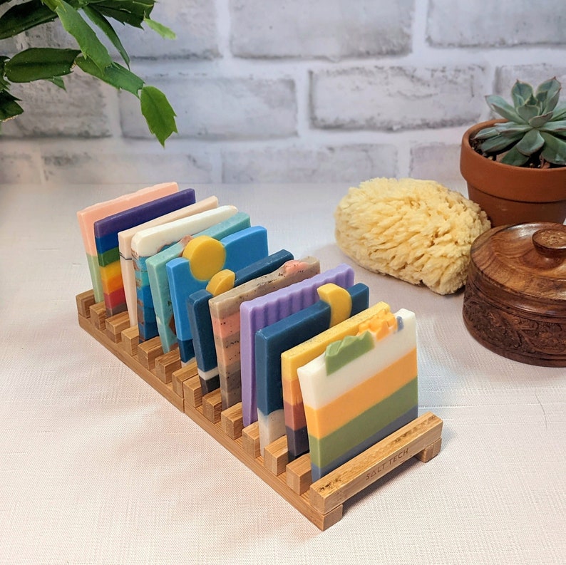 May include: A wooden soap holder displaying a variety of colorful, layered soap bars. The soaps feature shades of blue, green, yellow, orange, and purple. The holder is made of light brown wood and is labeled "SALT TECH".