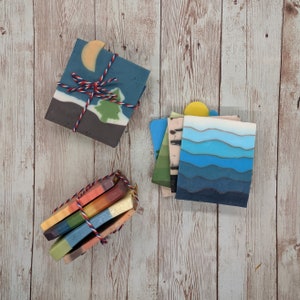 May include: A collection of handmade soaps with various designs. One soap features a night sky scene with a moon and tree. Another has blue water-like layers. A third set is a rainbow of colors, all tied with red, white, and blue twine.