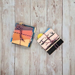 May include: Two stacks of handmade soap bars tied with red, white, and blue twine. One stack features layers of orange, pink, and purple. The other has pink, black, and white marbled designs. A yellow soap is on top.