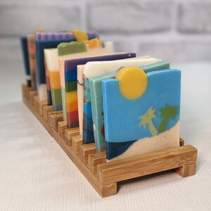 May include: A wooden soap holder displaying a variety of colorful, square-shaped soaps. The soaps feature unique designs, including a beach scene with a yellow sun and palm trees. The holder is made of light brown wood.