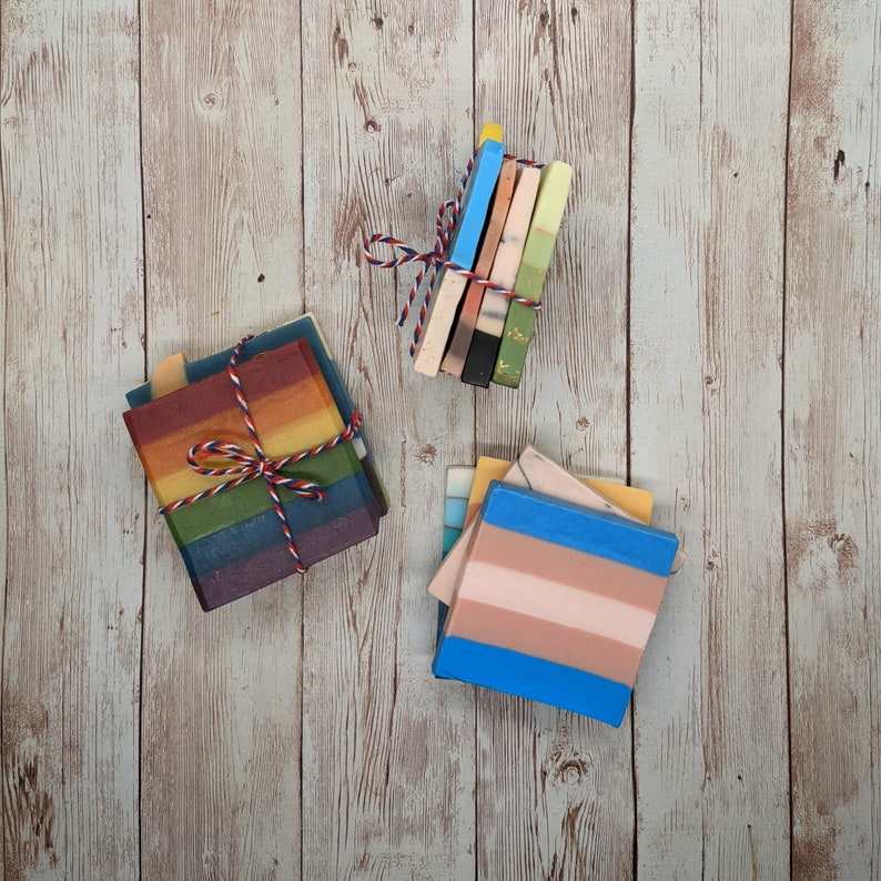 May include: Bundles of colorful handmade soaps tied with red, white, and blue twine. The soaps feature various designs, including rainbow stripes and color blocks. The soaps are arranged on a light wood-grain surface.
