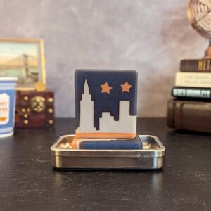 May include: A blue and orange bar of soap with a white silhouette of a city skyline and two stars. The soap is on a silver soap dish.