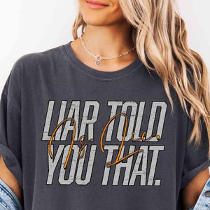 May include: A charcoal gray short-sleeve t-shirt featuring the text LIAR TOLD YOU THAT in a distressed white block font with the words Is Lie written in a cursive orange script overlaying the center of the graphic design.