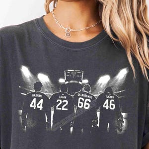 Peut inclure: Un t-shirt gris anthracite avec une impression graphique représentant quatre silhouettes en maillots de sport dans un stade. Les maillots portent les noms Graham, Logan, Di Laurentis et Tucker avec les numéros 44, 22, 66 et 46.