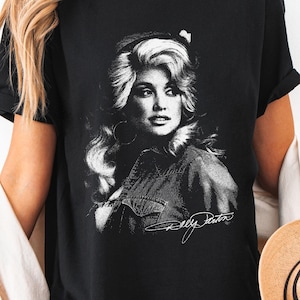 May include: Black t-shirt featuring a black and white portrait of a woman with long, wavy hair and a denim shirt. The text "Dolly Parton" is below the portrait. The shirt is a Gildan 64000.