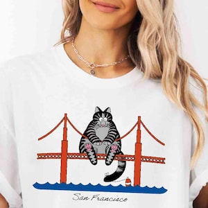 May include: A white t-shirt featuring a graphic of a black and gray striped cat sitting on a red Golden Gate Bridge structure above blue water, with the text San Francisco written in black cursive script below the bridge illustration.
