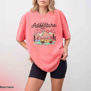 May include: A coral-coloured t-shirt featuring the text "And so the Adventure Begins" above a graphic of a yellow jeep with a kayak, mountains, trees, and a setting sun. The shirt has a loose fit.