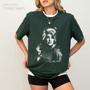May include: Forest green t-shirt featuring a black and white portrait of a woman with blonde hair. The shirt has a white collar and the text "Gildan 64000 Forest Green" is in the upper left corner. The woman's signature is at the bottom of the portrait.
