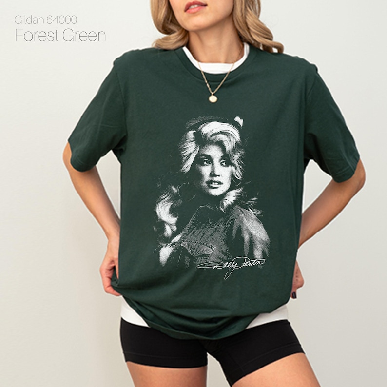May include: Forest green t-shirt featuring a black and white portrait of a woman with blonde hair. The shirt has a white collar and the text "Gildan 64000 Forest Green" is in the upper left corner. The woman's signature is at the bottom of the portrait.