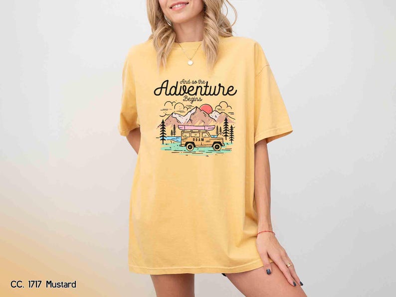 May include: Mustard yellow t-shirt featuring the text "And so the Adventure Begins" above a graphic of a vehicle with a kayak, mountains, and a rising sun. The shirt has a relaxed fit.
