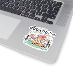 May include: A sticker depicting a yellow off-road vehicle with a pink surfboard on its roof, set against a mountain backdrop. The text "And so the Adventure Begins" is written above the scene.
