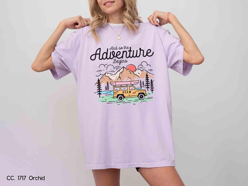 May include: A lilac t-shirt featuring the phrase "And so the Adventure Begins" above a graphic of a yellow vehicle with a kayak, mountains, trees, and a sunset. The shirt has a loose fit.