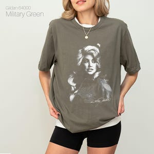 May include: Military green t-shirt featuring a black and white portrait of a celebrity with signature. The shirt has a crew neck and short sleeves. The text "Gildan 64000 Military Green" is printed on the upper left.