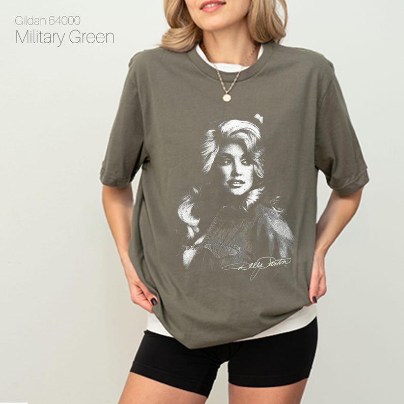 May include: Military green t-shirt featuring a black and white portrait of a celebrity with signature. The shirt has a crew neck and short sleeves. The text "Gildan 64000 Military Green" is printed on the upper left.