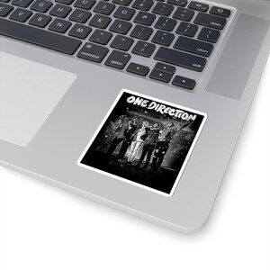 May include: A black and white sticker featuring the text "ONE DIRECTION" at the top. The sticker displays a group of five people in a band, standing together. The sticker is placed on a silver laptop.
