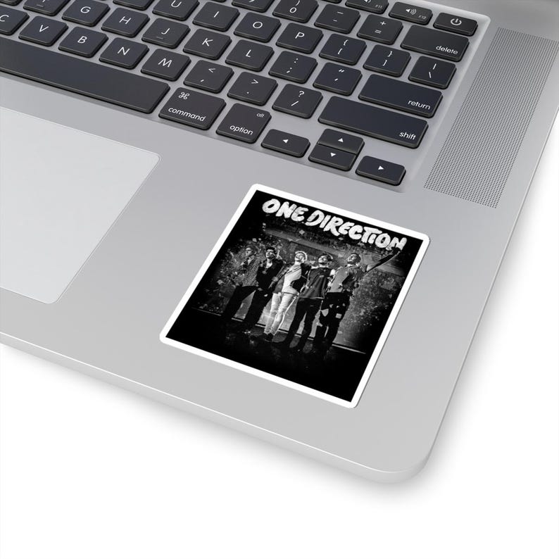 May include: A black and white sticker featuring the text "ONE DIRECTION" at the top. The sticker displays a group of five people in a band, standing together. The sticker is placed on a silver laptop.