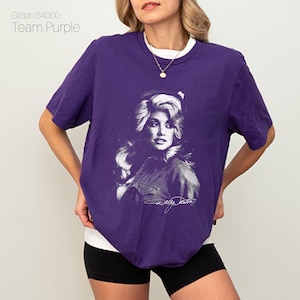 May include: A purple t-shirt with a portrait of a woman in black and white. The shirt has a white collar and the text "Gildan 64000 Team Purple" at the top left. The woman's signature is at the bottom of the portrait.
