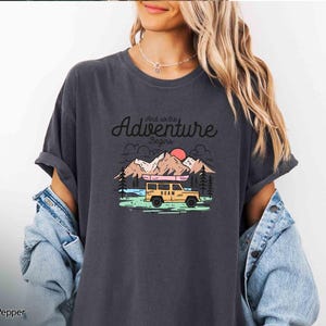 May include: Dark grey t-shirt featuring the text "And so the Adventure Begins" above a graphic of a yellow vehicle with a canoe, mountains, trees, and a setting sun. The shirt has a relaxed fit.