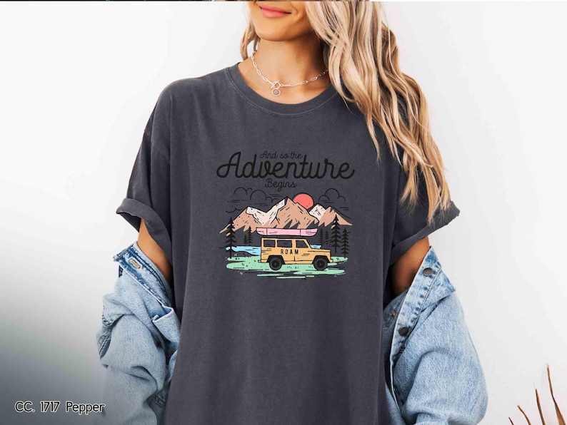 May include: Dark grey t-shirt featuring the text "And so the Adventure Begins" above a graphic of a yellow vehicle with a canoe, mountains, trees, and a setting sun. The shirt has a relaxed fit.