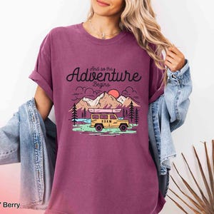 May include: Berry-coloured t-shirt featuring the text "And so the Adventure Begins" and a graphic of a yellow vehicle with a canoe on top, mountains, trees, and a sun. The shirt has rolled-up sleeves.