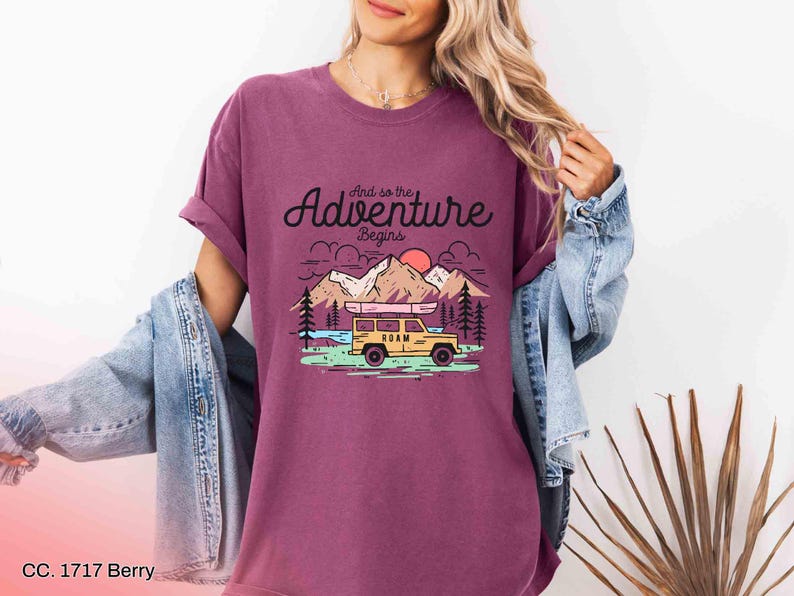 May include: Berry-coloured t-shirt featuring the text "And so the Adventure Begins" and a graphic of a yellow vehicle with a canoe on top, mountains, trees, and a sun. The shirt has rolled-up sleeves.