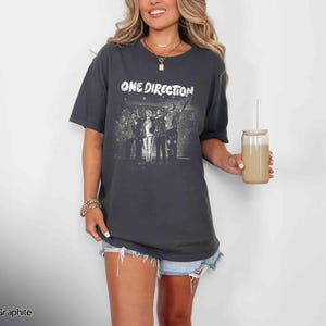 May include: Graphite gray t-shirt featuring the text "ONE DIRECTION" above a black and white image of the band. The shirt is worn with denim shorts. A glass with a straw is held in the hand.
