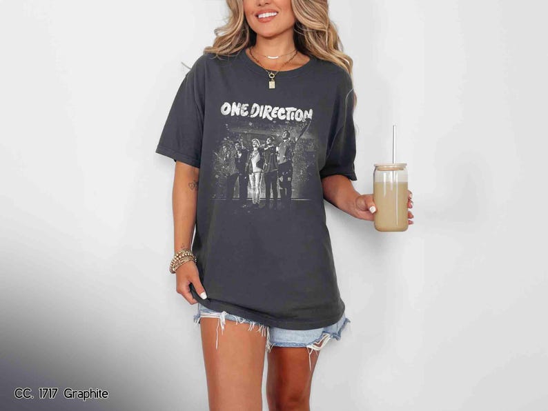 May include: Graphite gray t-shirt featuring the text "ONE DIRECTION" above a black and white image of the band. The shirt is worn with denim shorts. A glass with a straw is held in the hand.