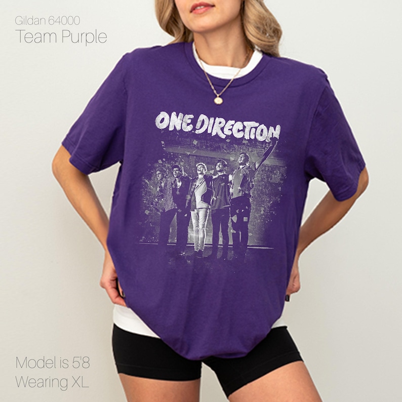 May include: A purple t-shirt with the text "ONE DIRECTION" in white, featuring a band image. The shirt is a Gildan 64000 in Team Purple. The model is 5'8 and wearing XL.