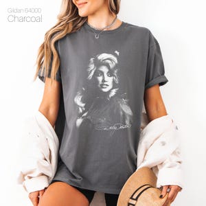 May include: Charcoal gray t-shirt featuring a black and white portrait of a woman with long, wavy hair and a signature. The text "Gildan 64000" is printed at the top left. The shirt is a comfortable, casual style.