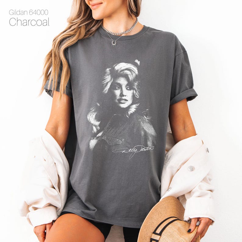 May include: Charcoal gray t-shirt featuring a black and white portrait of a woman with long, wavy hair and a signature. The text "Gildan 64000" is printed at the top left. The shirt is a comfortable, casual style.