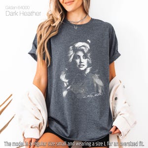 May include: Dark heather gray t-shirt featuring a black and white portrait of a woman with long hair and a signature. The shirt is labeled "Gildan 64000" and the text "Dark Heather" is visible. The model is wearing a size L for an oversized fit.