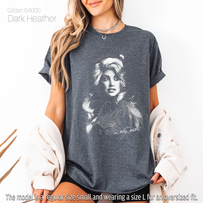May include: Dark heather gray t-shirt featuring a black and white portrait of a woman with long hair and a signature. The shirt is labeled "Gildan 64000" and the text "Dark Heather" is visible. The model is wearing a size L for an oversized fit.