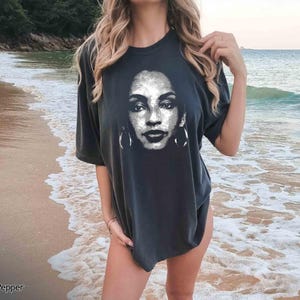 Peut inclure: Un t-shirt noir avec un imprim&eacute; graphique d'un visage de femme. La femme a les cheveux fonc&eacute;s et porte des boucles d'oreilles. Le t-shirt est port&eacute; par une personne debout sur une plage.
