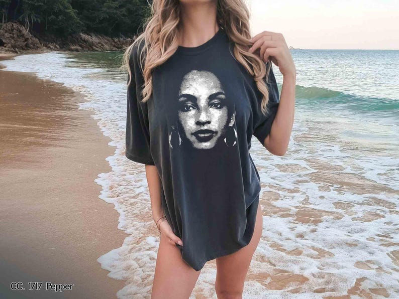 Peut inclure: Un t-shirt noir avec un imprim&eacute; graphique d'un visage de femme. La femme a les cheveux fonc&eacute;s et porte des boucles d'oreilles. Le t-shirt est port&eacute; par une personne debout sur une plage.