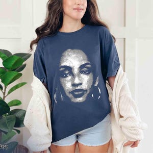 Peut inclure: Un t-shirt bleu marine avec un motif blanc repr&eacute;sentant le visage d'une femme. Le motif est un gros plan du visage de la femme, et il comprend ses yeux, son nez, sa bouche et ses boucles d'oreilles.
