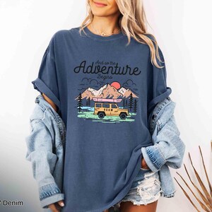 May include: A navy blue t-shirt featuring the phrase "And so the adventure begins" above a graphic of a yellow SUV with a kayak, mountains, trees, and a sunset. The shirt has a relaxed fit.