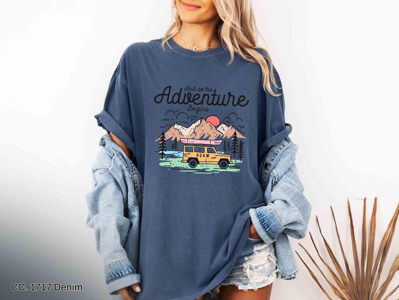 May include: A navy blue t-shirt featuring the phrase "And so the adventure begins" above a graphic of a yellow SUV with a kayak, mountains, trees, and a sunset. The shirt has a relaxed fit.