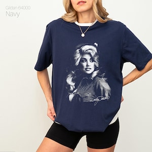 May include: Navy blue t-shirt featuring a black and white portrait of a celebrity with signature. The shirt has a white collar and the text "Gildan 64000 Navy" is printed on the upper left side.