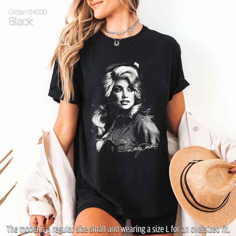 May include: Black t-shirt featuring a black and white portrait of a woman with long, wavy hair and a denim shirt. The text "Dolly Parton" is below the portrait. The shirt is a Gildan 64000.