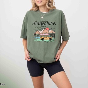 May include: An olive green t-shirt featuring the text "And so the Adventure begins" above a graphic of a yellow vehicle, mountains, and a red sun. The shirt is worn with black shorts.