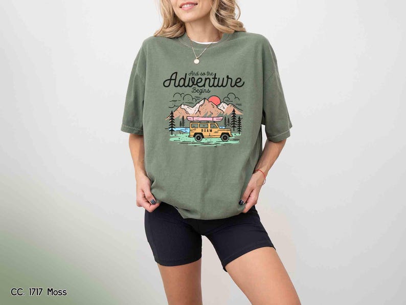 May include: An olive green t-shirt featuring the text "And so the Adventure begins" above a graphic of a yellow vehicle, mountains, and a red sun. The shirt is worn with black shorts.