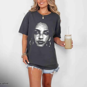Peut inclure: Un t-shirt gris fonc&eacute; avec un motif blanc repr&eacute;sentant le visage d'une femme. La femme porte de grandes boucles d'oreilles cr&eacute;oles. Le t-shirt est port&eacute; par une personne aux cheveux longs et bruns.