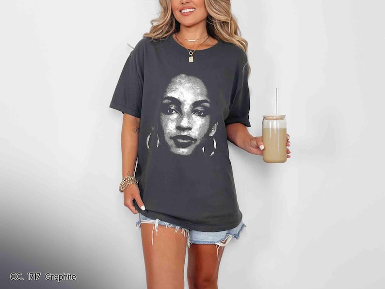 Peut inclure: Un t-shirt gris fonc&eacute; avec un motif blanc repr&eacute;sentant le visage d'une femme. La femme porte de grandes boucles d'oreilles cr&eacute;oles. Le t-shirt est port&eacute; par une personne aux cheveux longs et bruns.
