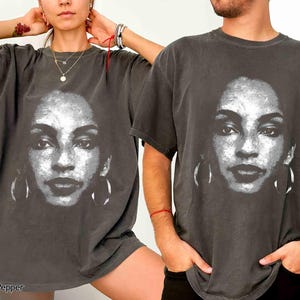 Peut inclure: Un t-shirt noir avec un imprim&eacute; graphique blanc d'un visage de femme. Le graphique est un gros plan du visage de la femme avec de grandes boucles d'oreilles. Le t-shirt est port&eacute; par deux mannequins, une femme et un homme.