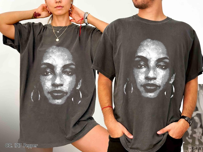 Peut inclure: Un t-shirt noir avec un imprim&eacute; graphique blanc d'un visage de femme. Le graphique est un gros plan du visage de la femme avec de grandes boucles d'oreilles. Le t-shirt est port&eacute; par deux mannequins, une femme et un homme.