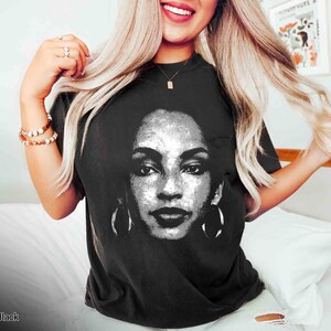 Peut inclure: Un t-shirt noir avec une image blanche de la chanteuse Aaliyah imprim&eacute;e sur le devant. L'image est un gros plan de son visage avec de grandes boucles d'oreilles.
