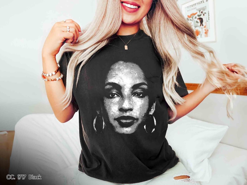 Peut inclure: Un t-shirt noir avec une image blanche de la chanteuse Aaliyah imprim&eacute;e sur le devant. L'image est un gros plan de son visage avec de grandes boucles d'oreilles.